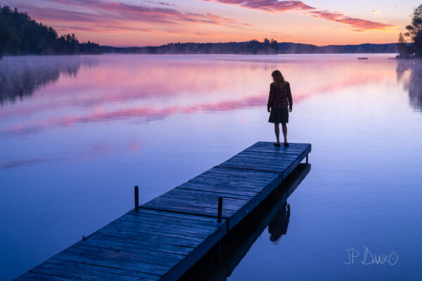 Woman (43) on cottage dock with colorful summer sunrise.
